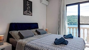 A bedroom with a bed, a nightstand, a wall-mounted air conditioner, and a window with a view of a lake and hills. - Apartments Mila (Ubli)