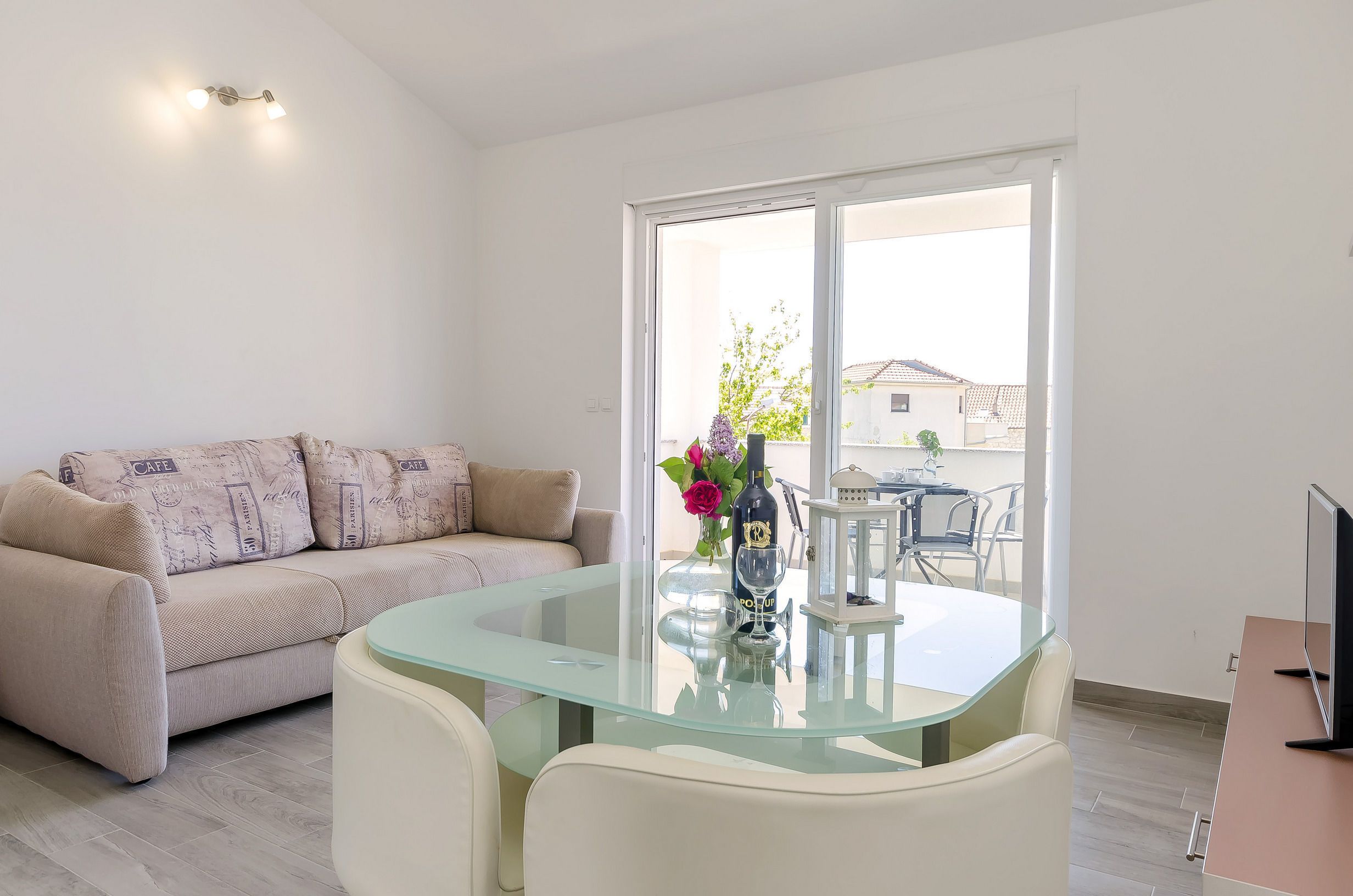 A modern living room with a glass-top dining table, a beige sofa with patterned cushions, a vase with flowers, and a view of the outside through a large sliding glass door.