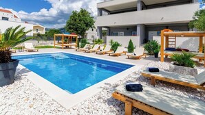A modern outdoor pool area with lounge chairs and a wooden pergola, surrounded by white buildings and greenery. - Apartments and Room Gabriels Paradise (Kastel Luksic)