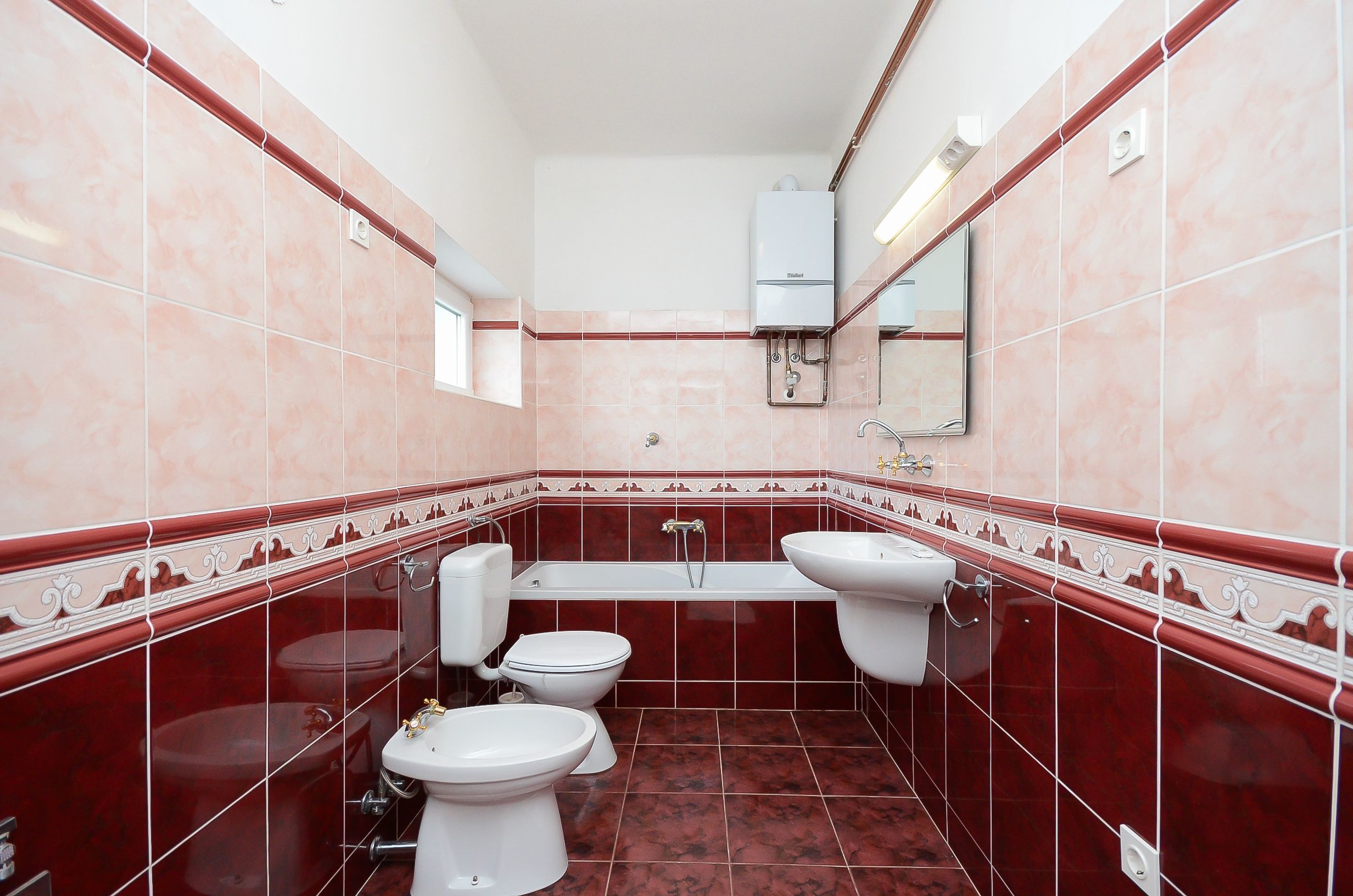 Een badkamer met toilet, bidet en wastafel, alles wit, met rode betegelde muren en een wit plafond.