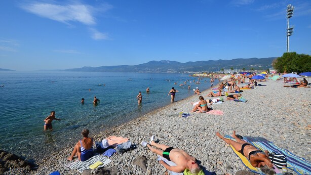 Ein Kiesstrand mit badenden und sonnenden Menschen im Wasser.