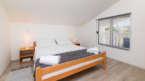 A bedroom with a wooden bed, white bedding, a nightstand with a lamp, a window with blinds, and a view of greenery outside. - Apartment Loro (Ploce)