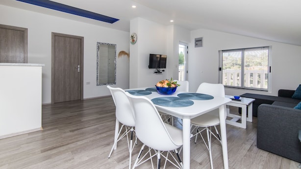 A modern living room with a white dining table, white chairs, a grey sofa, and a TV mounted on the wall. - Apartment Loro (Ploce)