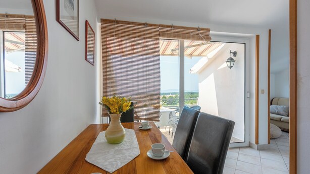 A dining area with a wooden table, two chairs, a vase with yellow flowers, and a view of the outdoors through a glass door. - Apartment Iva (Banjol)