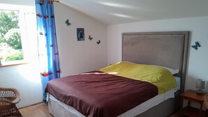 A bedroom with a bed, a window with curtains, a bedside table with a lamp, and a wicker chair. - Apartment Iva (Banjol)