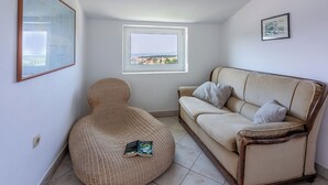 A room with a beige sofa, a wicker chair, a framed picture, and a window with a view of buildings. - Apartment Iva (Banjol)