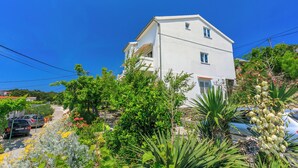 A two-story white building with a balcony, surrounded by lush greenery and flowering plants. - Apartment Iva (Banjol)