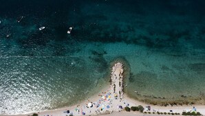 Vista aérea de uma praia com águas cristalinas, faixa de areia e um pequeno afloramento rochoso.