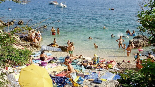 Una playa concurrida donde gente nada y toma el sol, con barcos en el agua y sombrillas sobre la arena.