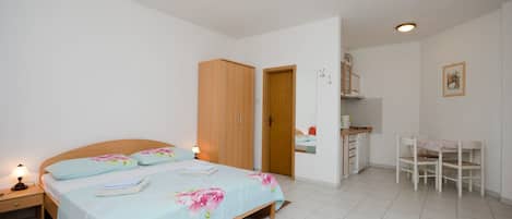 A bedroom with a bed, bedside table, wardrobe, desk, chair, and a small kitchenette.