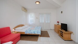 Un dormitorio con cama, un sofá rojo, un televisor sobre un soporte de madera y una puerta blanca con contraventanas.