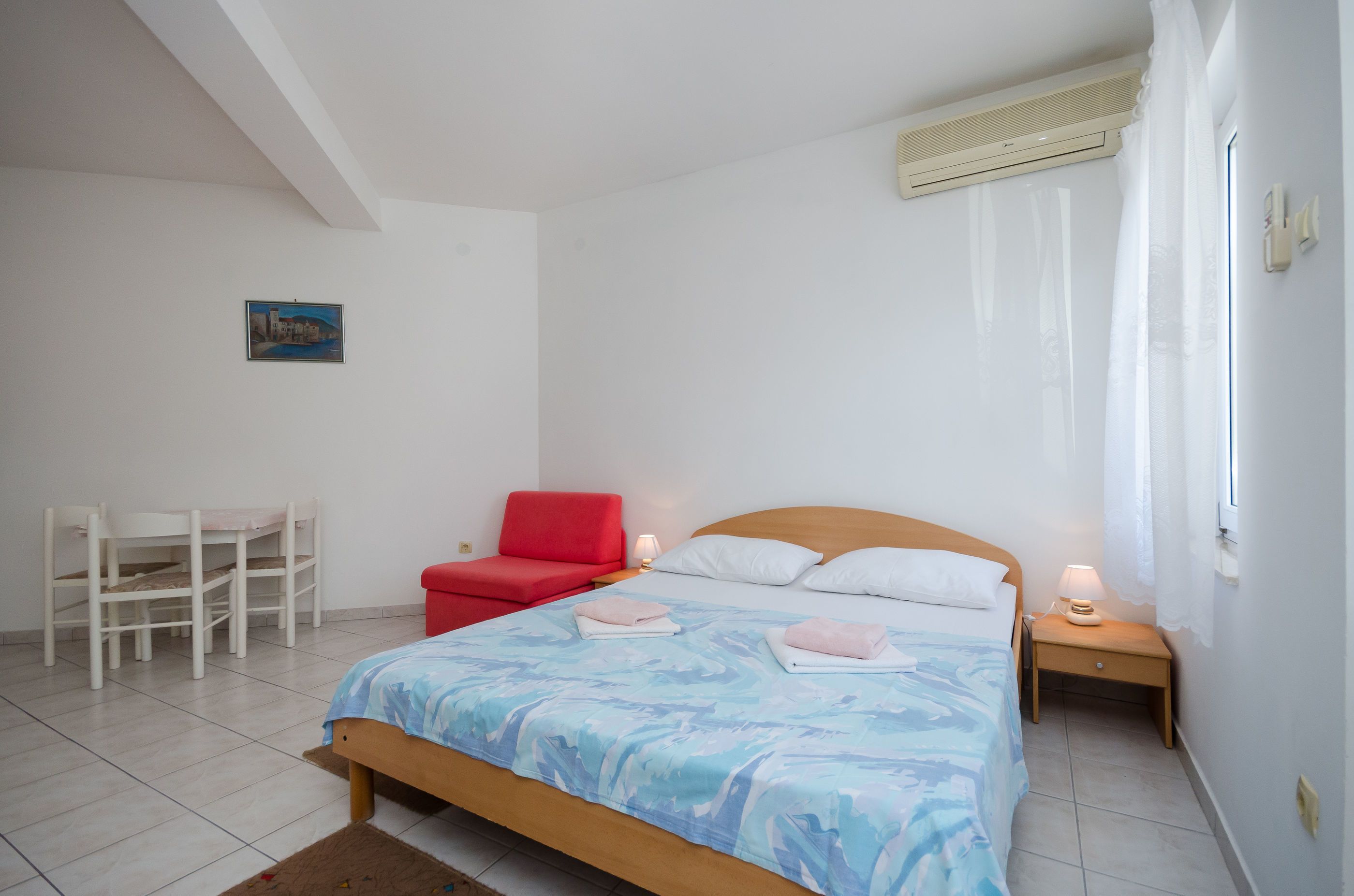 A bedroom with a bed, a red chair, a desk, and a nightstand with a lamp.