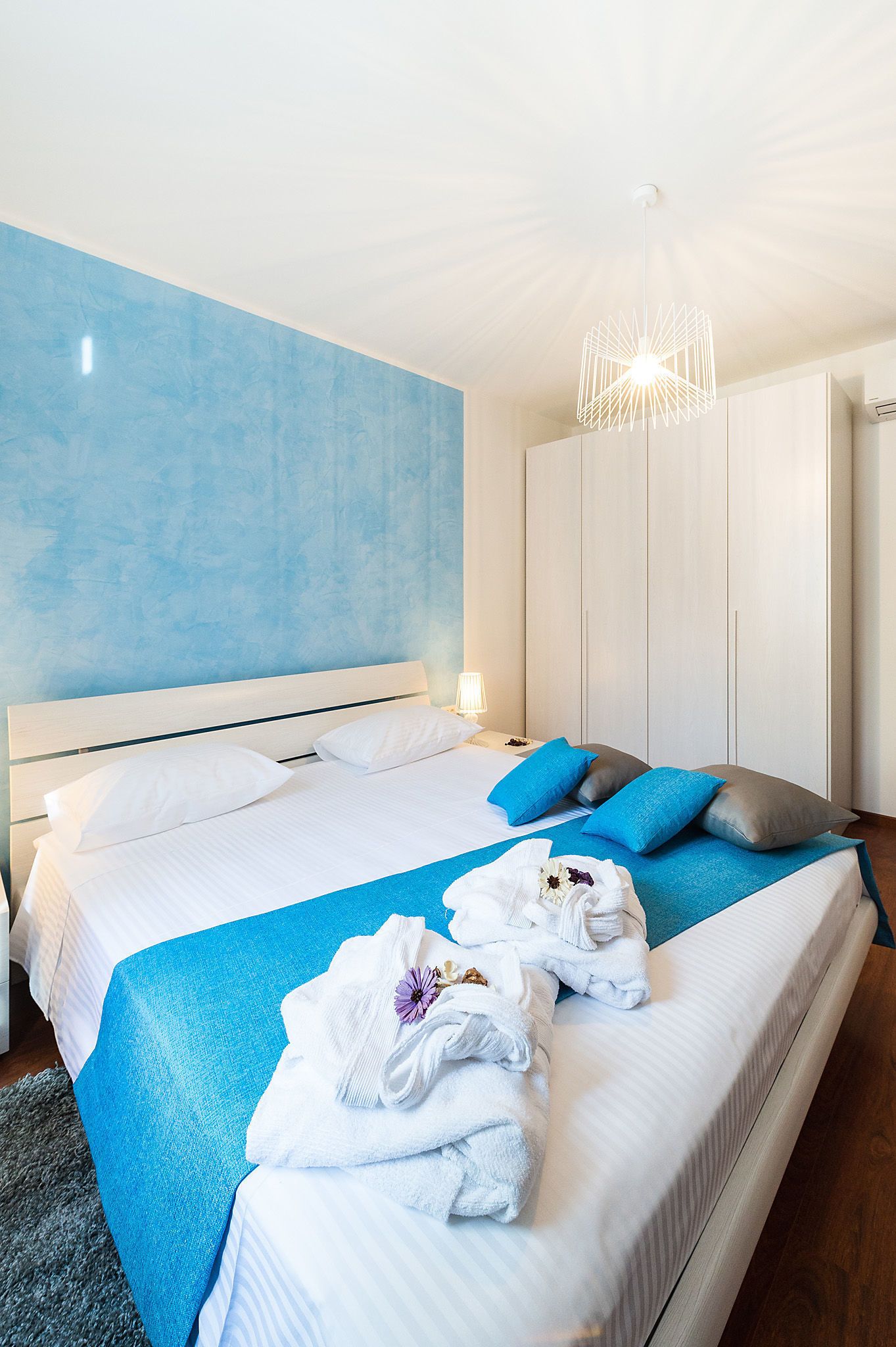 A hotel room with a large bed, a blue accent wall, a modern chandelier, and a wardrobe.