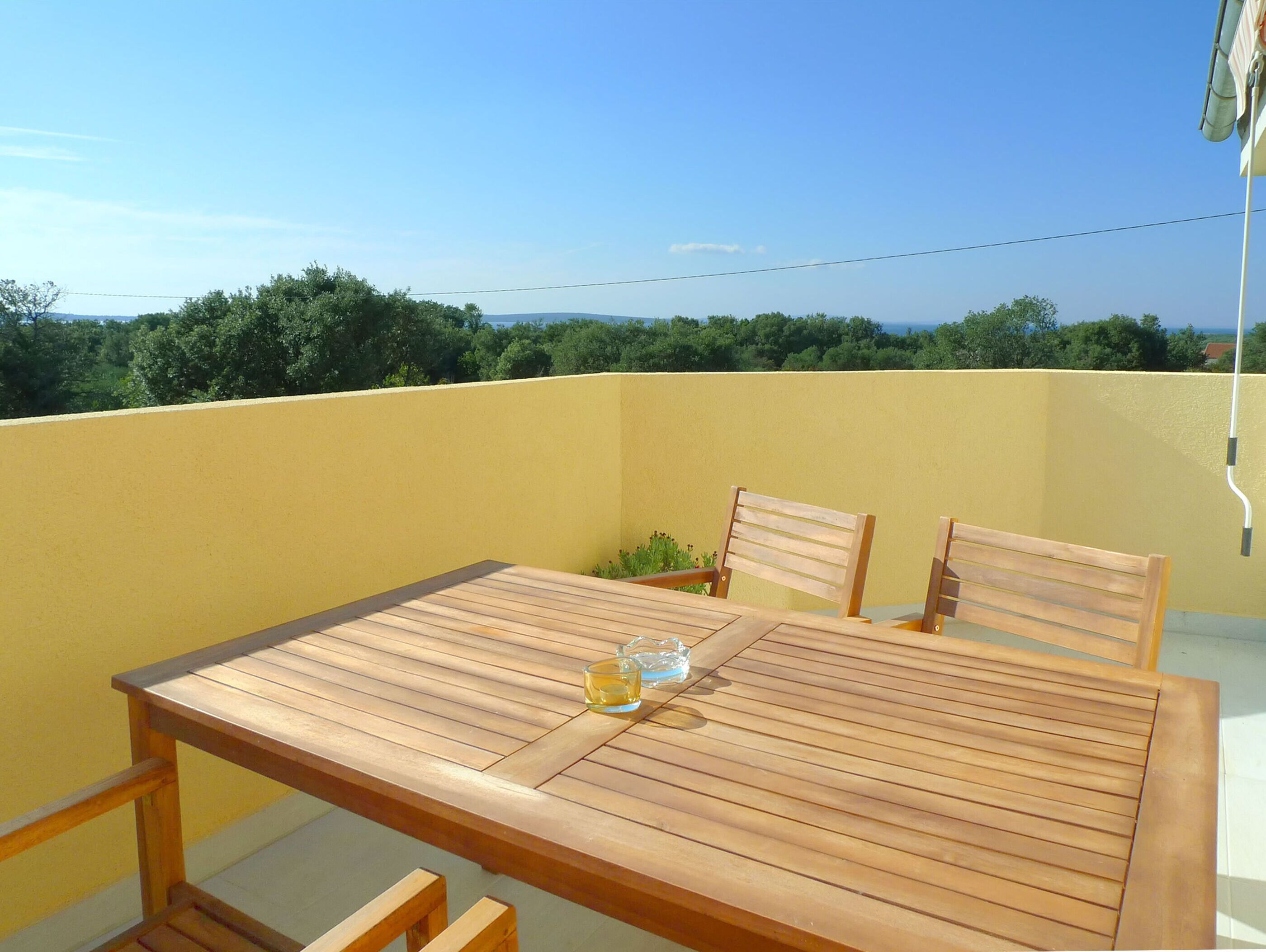 A wooden table with chairs on a balcony with a yellow wall and a view of trees and mountains.