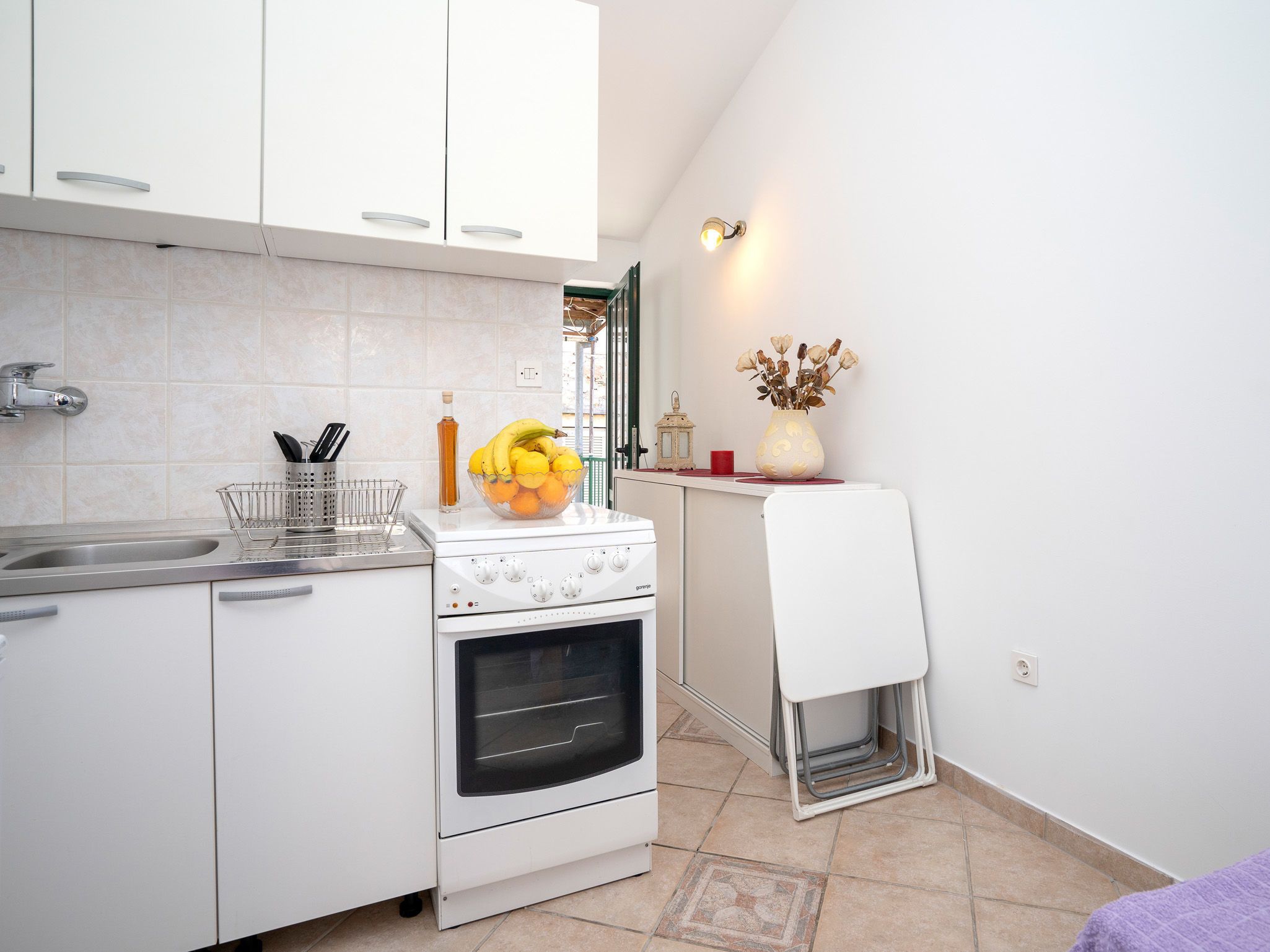 Una cocina compacta con armarios blancos, un fregadero, una estufa y una pequeña encimera con un cuenco de frutas y un jarrón.