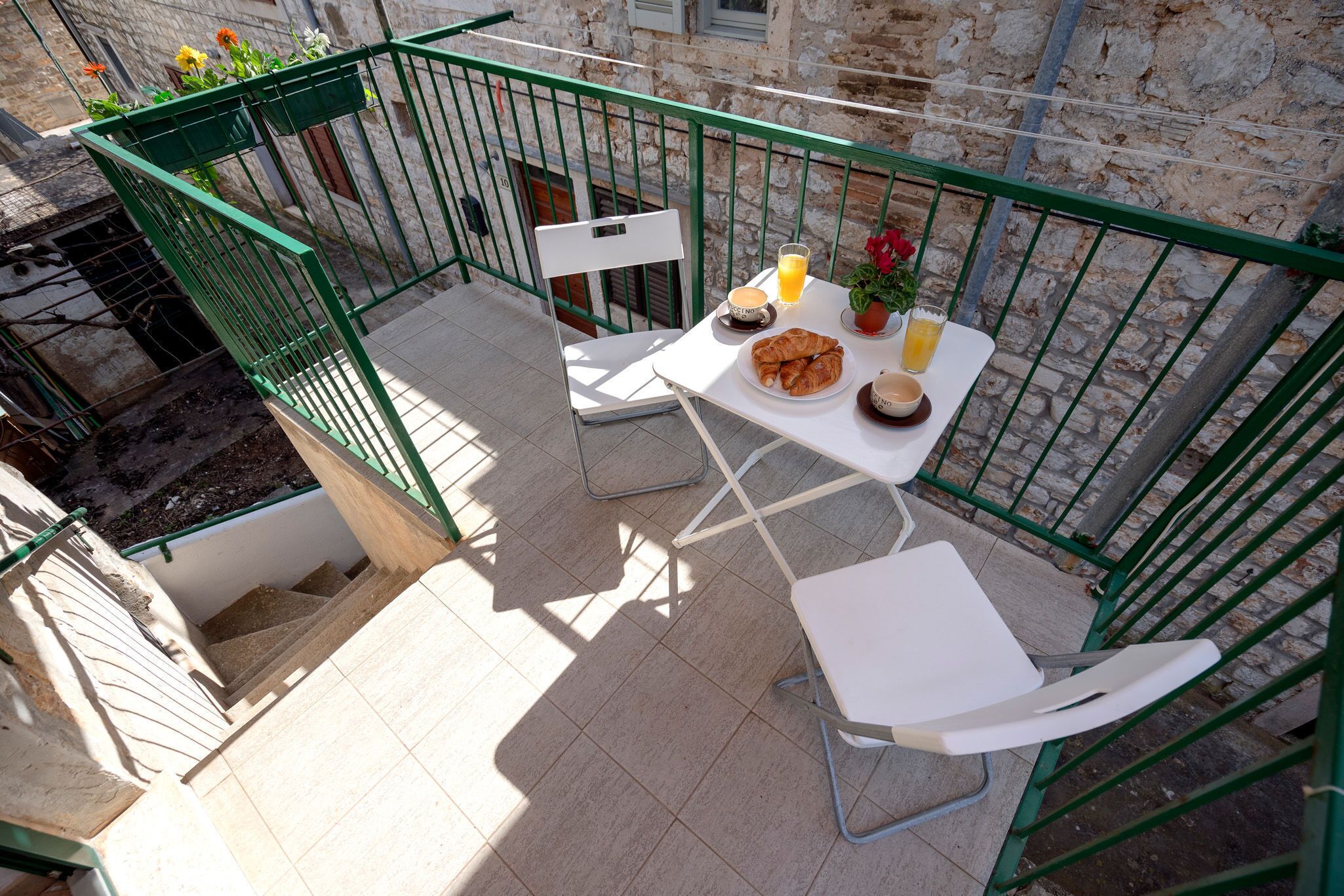 A balcony with a table set for breakfast, including croissants, coffee, and juice, with a view of a stone building.