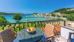 Un balcón con una mesa dispuesta con fruta, con vistas a un pueblo costero con casas y un cielo azul despejado.