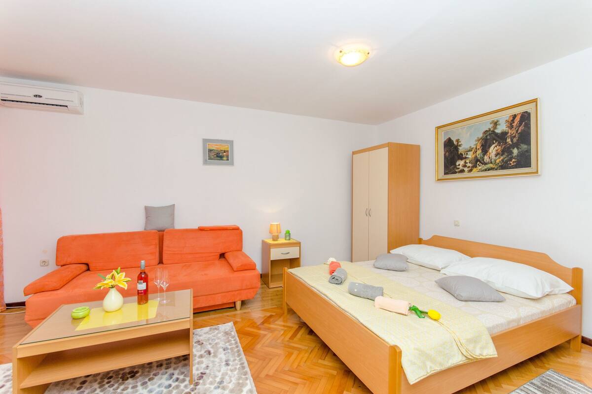 A bedroom with a bed, a nightstand, a red sofa, a wooden wardrobe, and a painting on the wall.