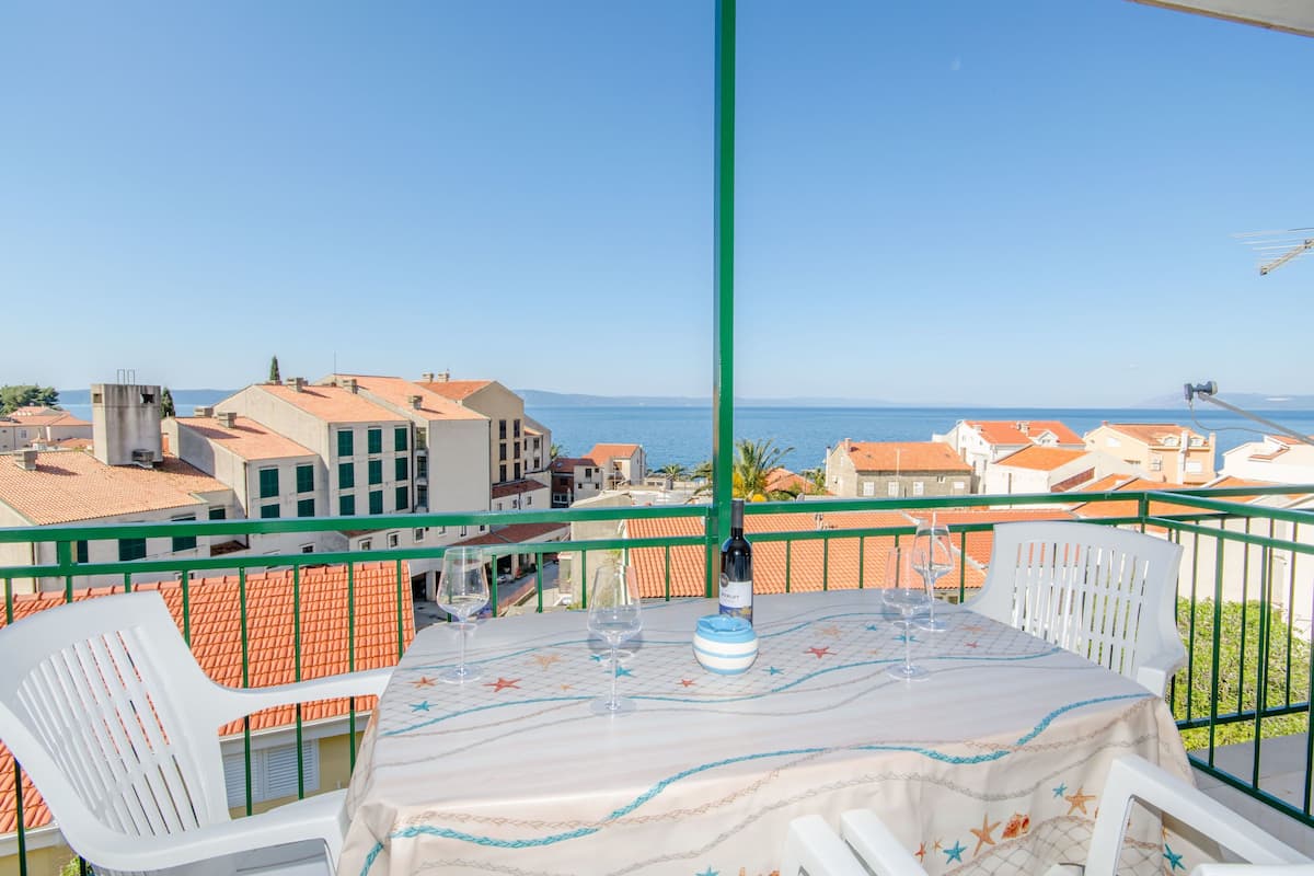 A balcony with a table set for two, overlooking a coastal town with terracotta-roofed buildings and a clear blue sea.