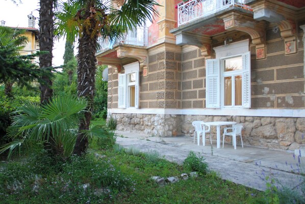 A building with stone walls, white shutters, and a balcony with white railings. There are two white chairs outside. - Apartments Rezine (Lovran)