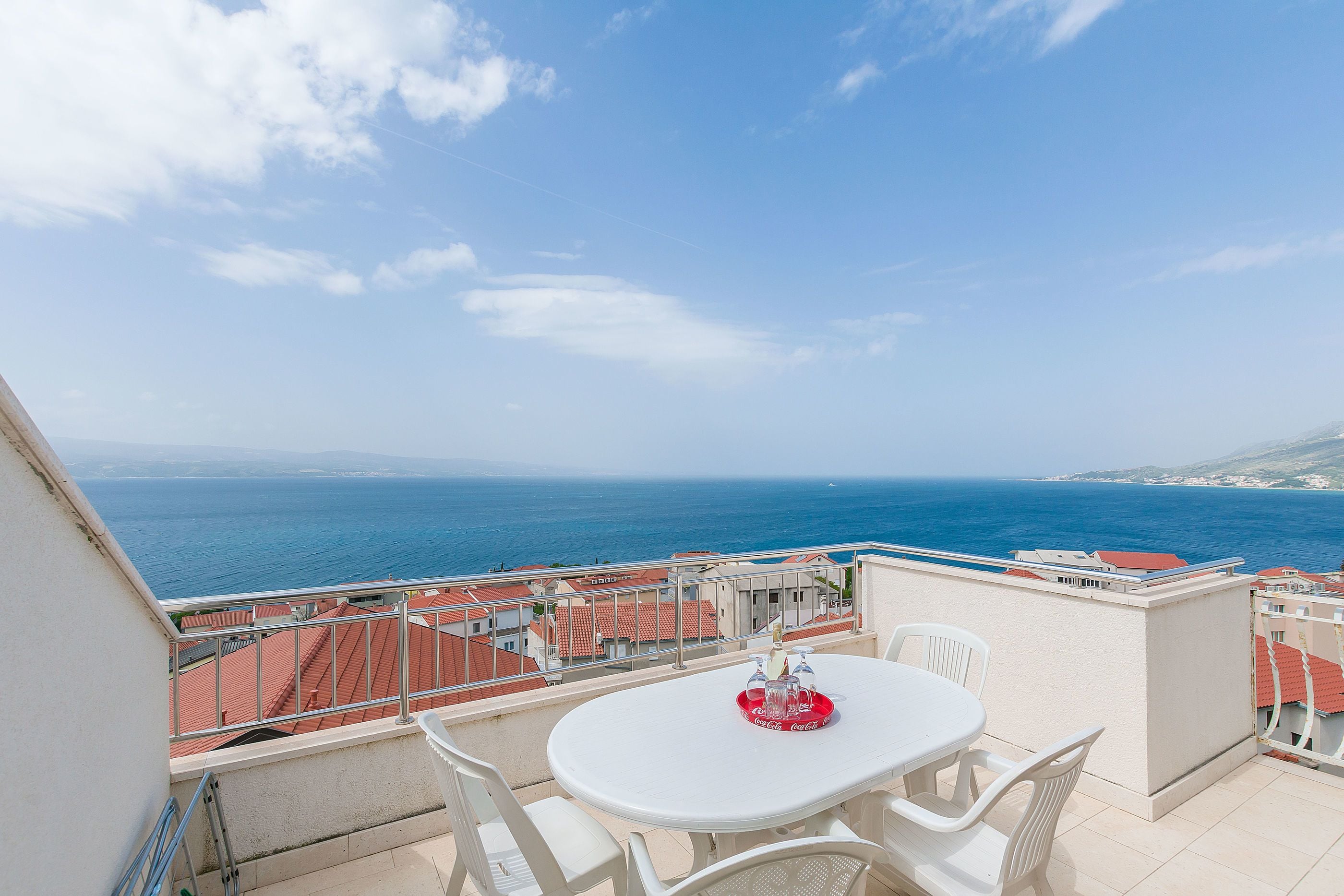 A balcony with a round table and chairs, overlooking a coastal view with buildings and the sea.