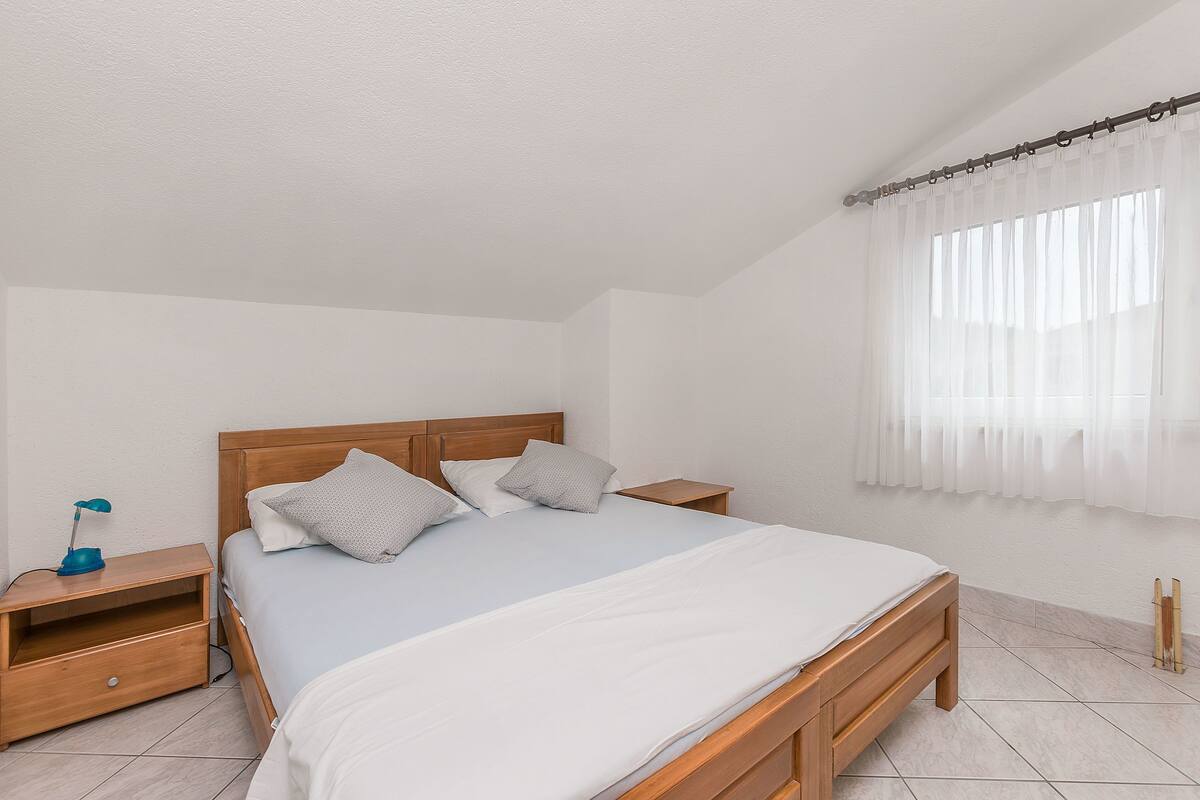 Un dormitorio con cama de madera, ropa de cama blanca, una mesita de noche con lámpara y una ventana con cortinas translúcidas.