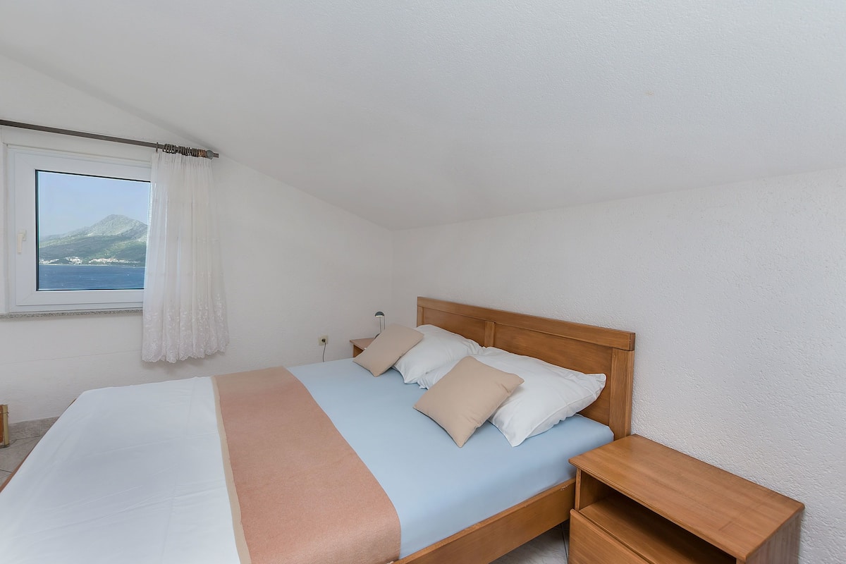 Un dormitorio con cama, mesita de noche de madera y ventana con vista al mar y a la montaña.