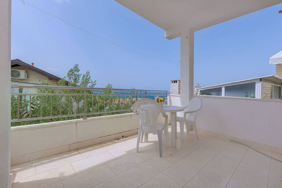 Un balcón con una silla y una mesa blancos, barandilla y vista al mar y edificios.