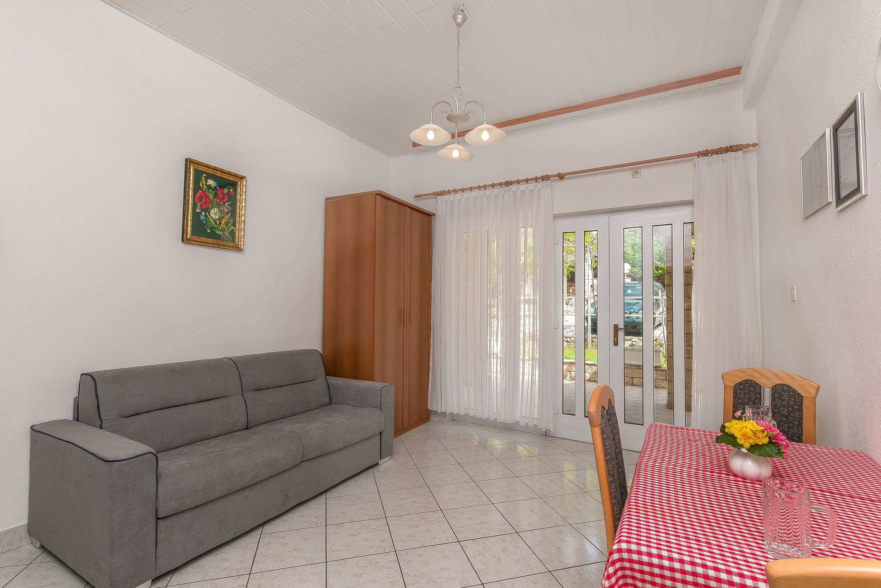 A living room with a grey sofa, a wooden wardrobe, a dining table with a red checkered tablecloth, and a glass pitcher.