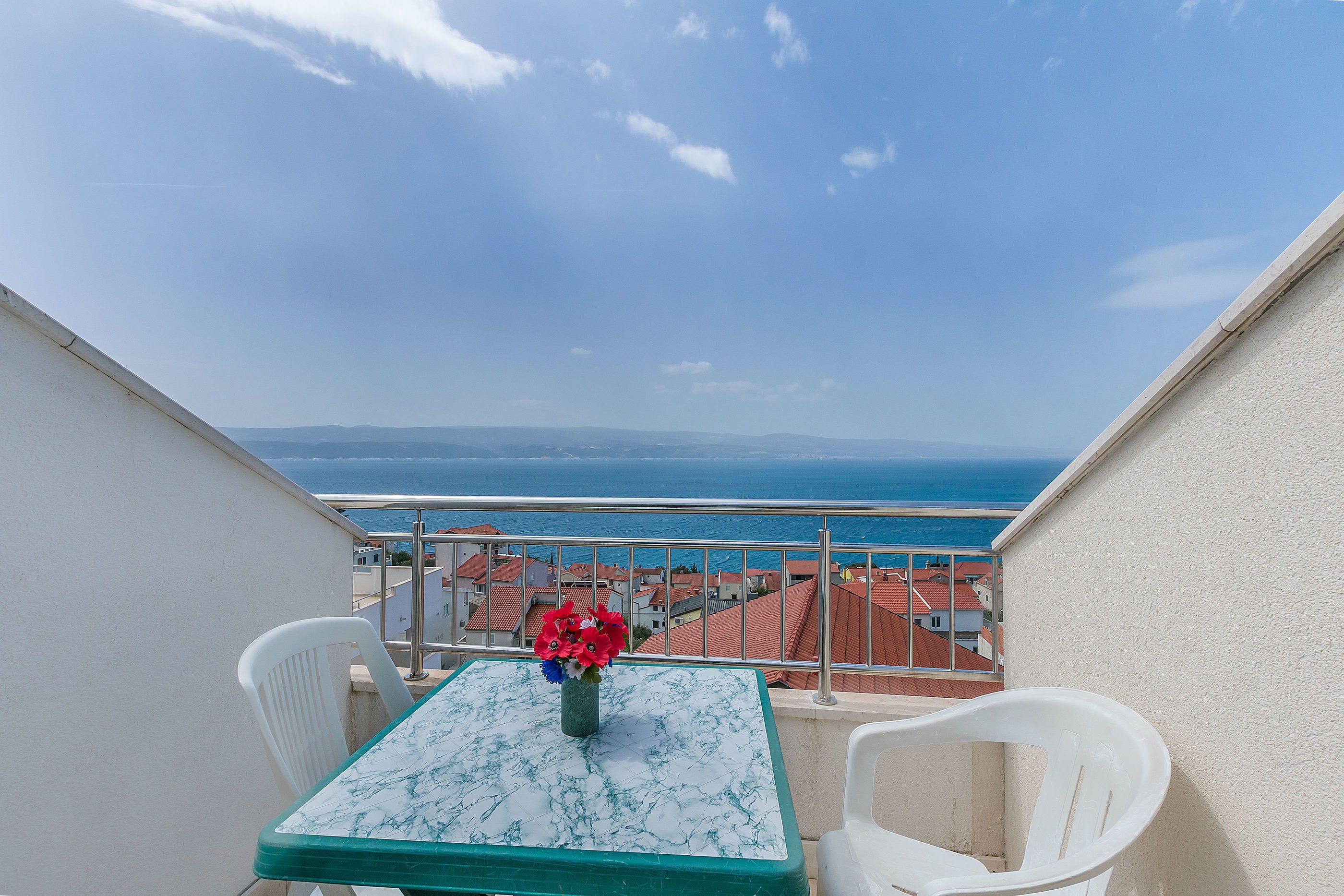 A balcony with a table and chairs, a vase with flowers, and a view of the sea and buildings.