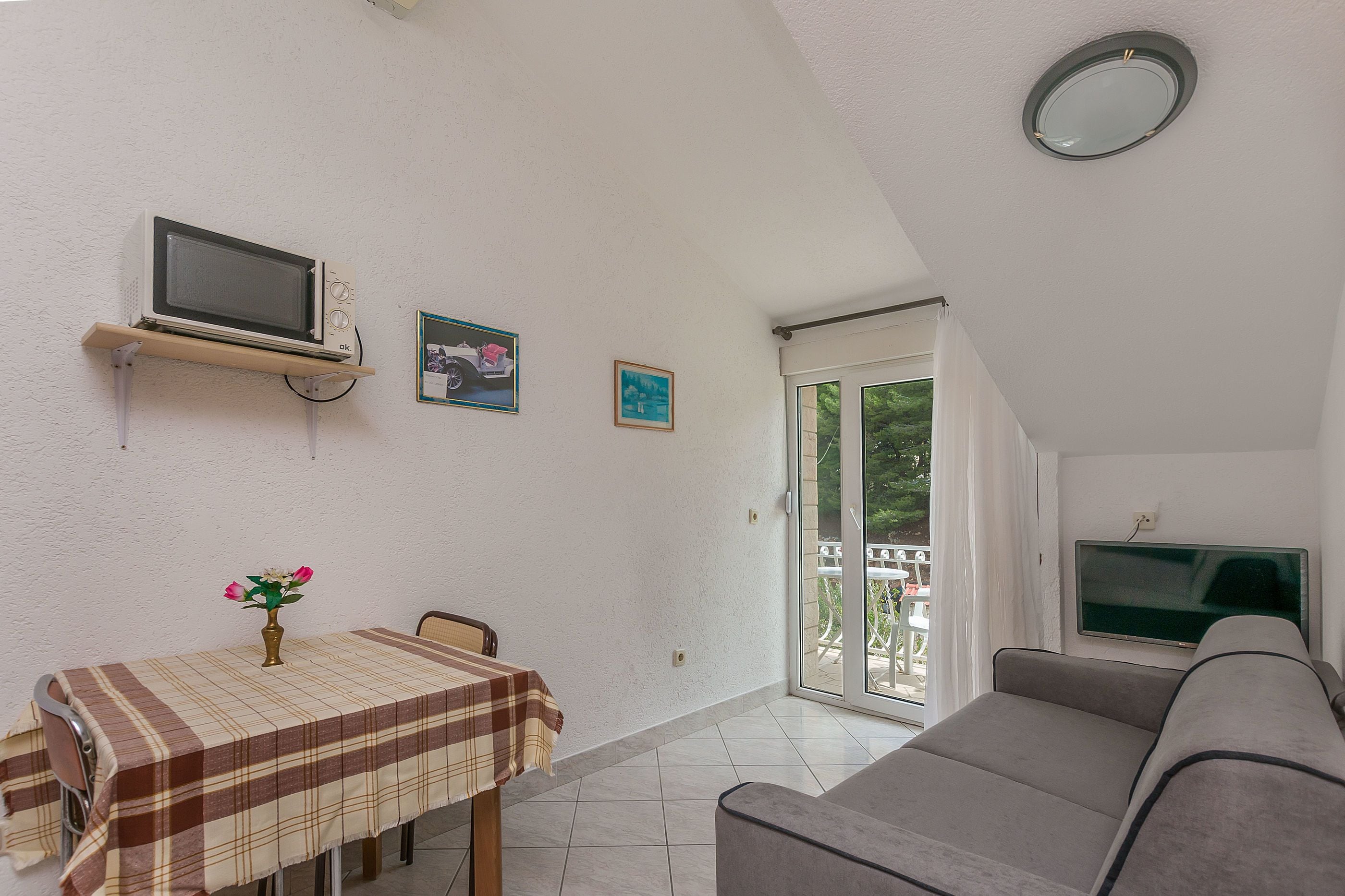 A room with a dining table, a sofa, a TV mounted on the wall, and a balcony door.