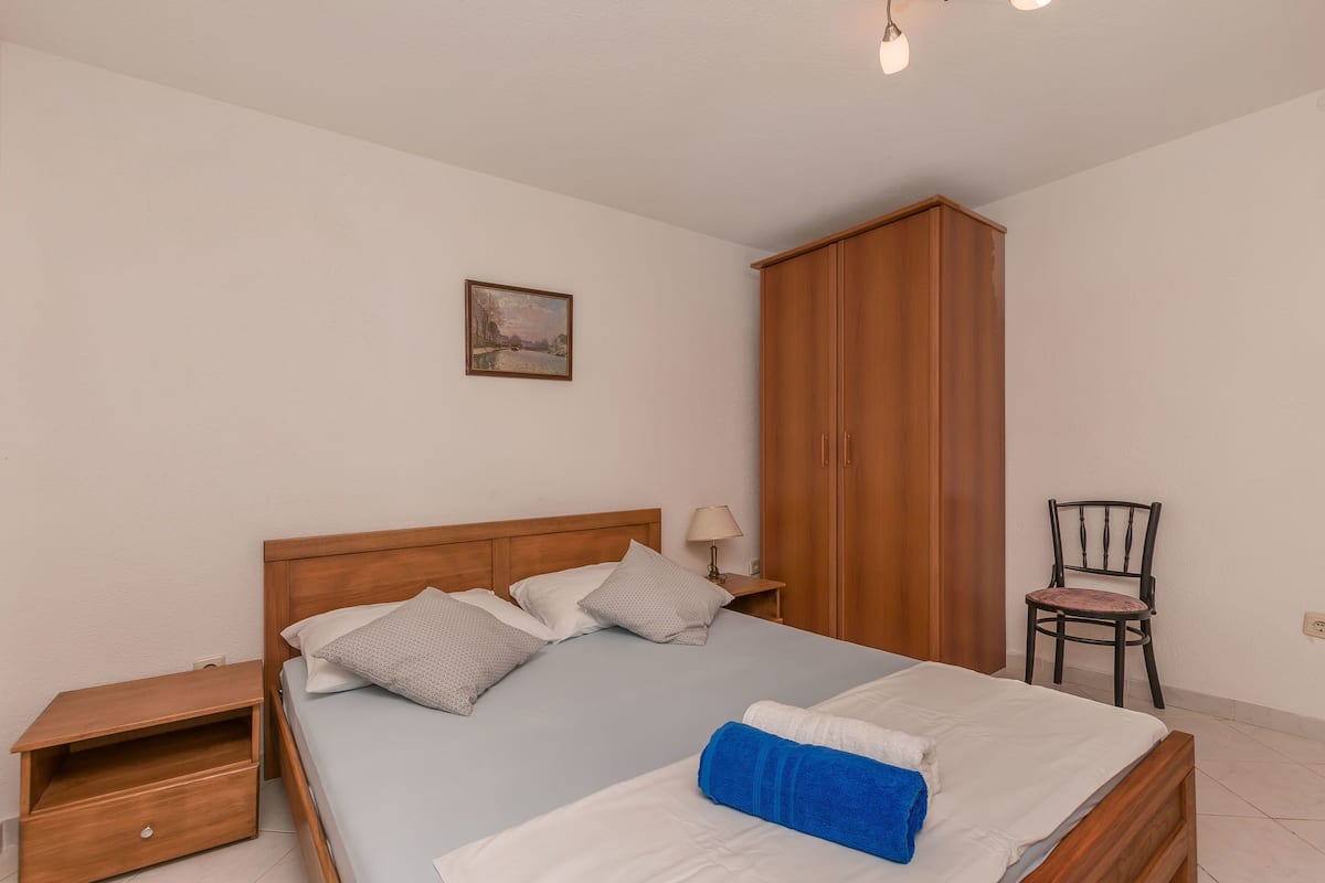 Una cama bien tendida con almohadas, un armario de madera, una silla y una mesita de noche con una lámpara.