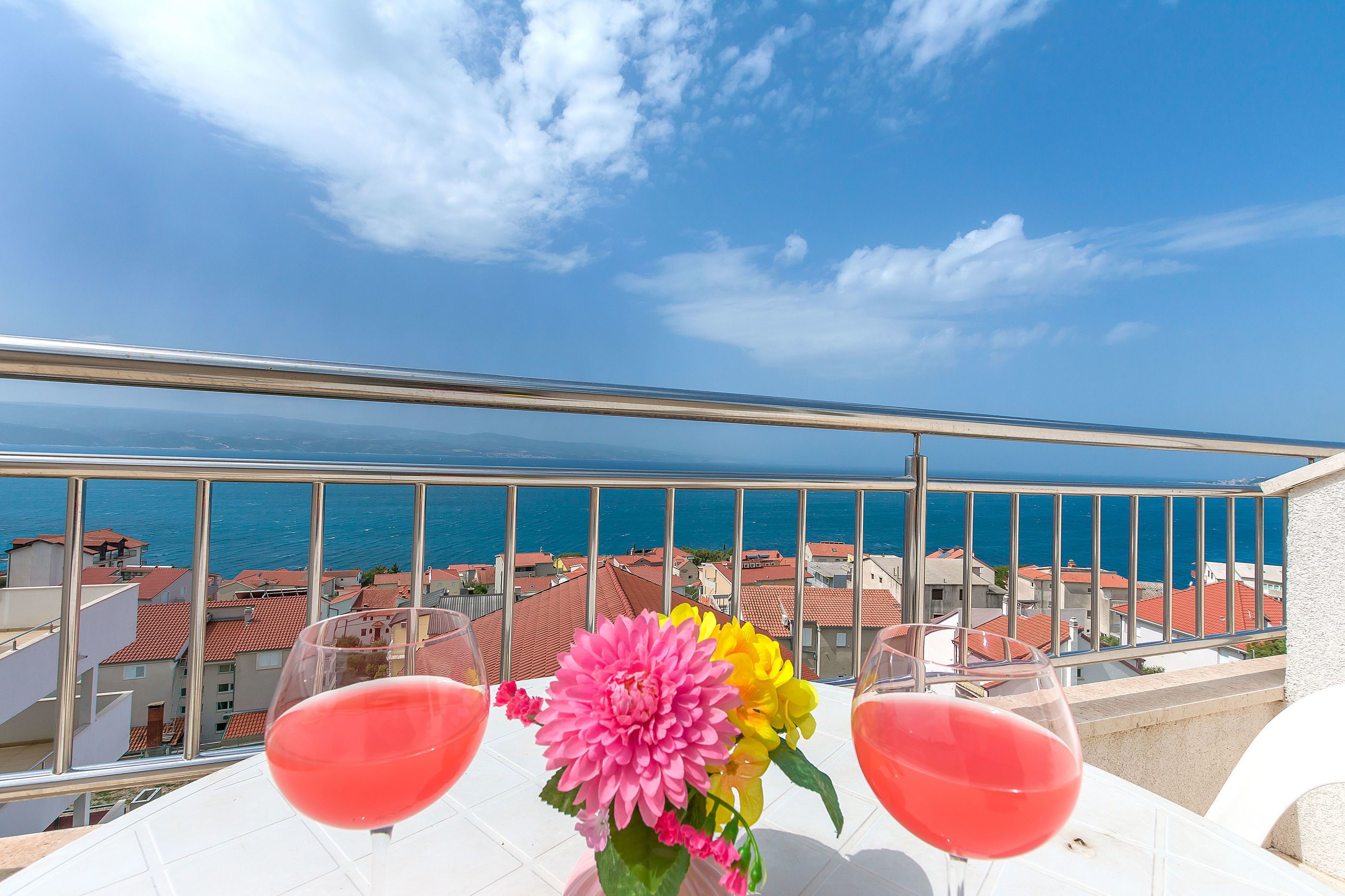 Ein Balkon mit zwei Weingläsern, einer rosa und gelben Blume und Blick auf das Meer und die Gebäude.
