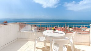 Ein Balkon mit einem weißen Tisch und Stühlen, einer Blumenvase sowie Blick auf das Meer und Gebäude.