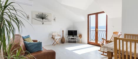 A spacious living area with a brown leather sofa, a wooden dining table with chairs, a television on a stand, and a large window with a view.