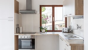 Une cuisine moderne équipée d’un four encastré, d’une plaque de cuisson et d’une hotte en acier inoxydable. Une fenêtre offre une vue sur un immeuble, et une plante en pot est posée sur le plan de travail.