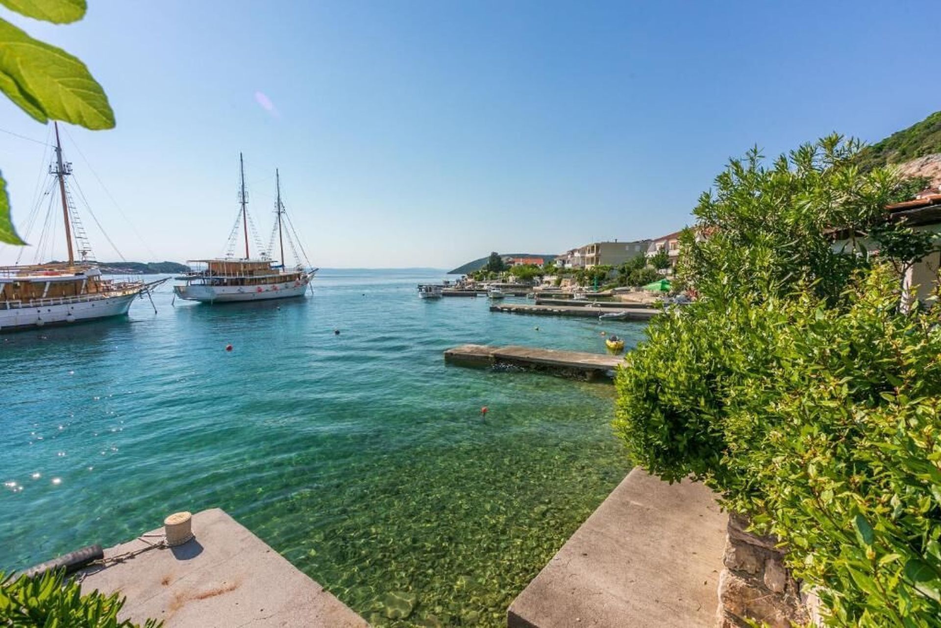 A coastal marina with boats, clear water, and a stone pathway leading to a building.