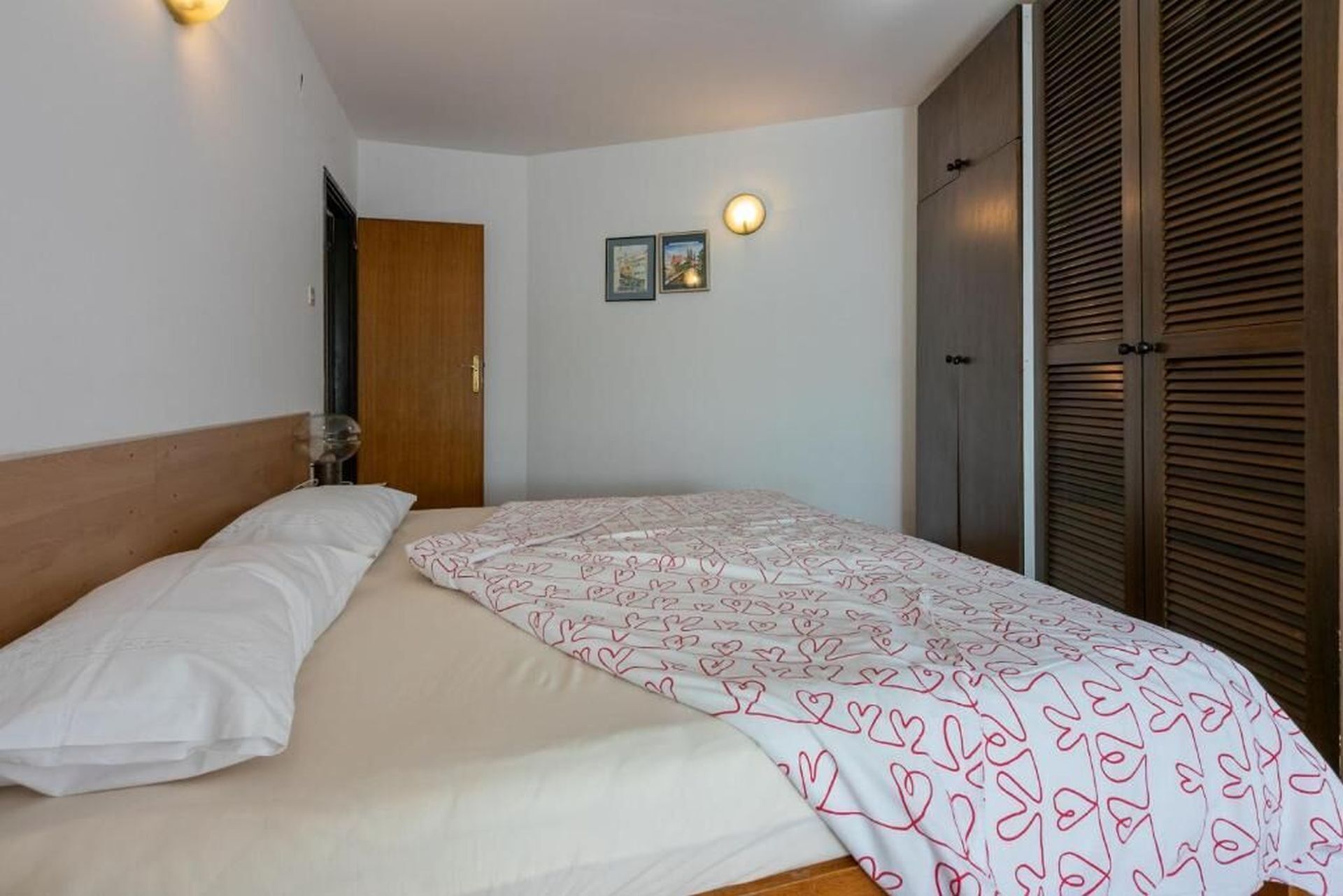 A bedroom with a bed, a wooden wardrobe, a door, and two wall-mounted light fixtures.