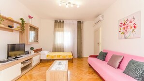 A modern living room with a wooden floor, a pink sofa, a TV, a small table, and a bed with a yellow bedspread. - Apartment Sime (Brodarica)