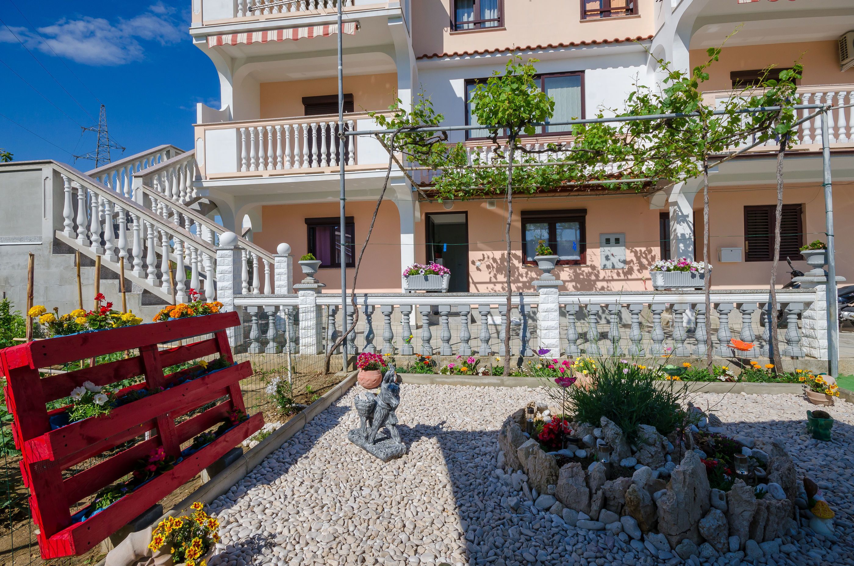A multi-story residential building with a balcony, a red bench, and a garden area with flowers and a statue.