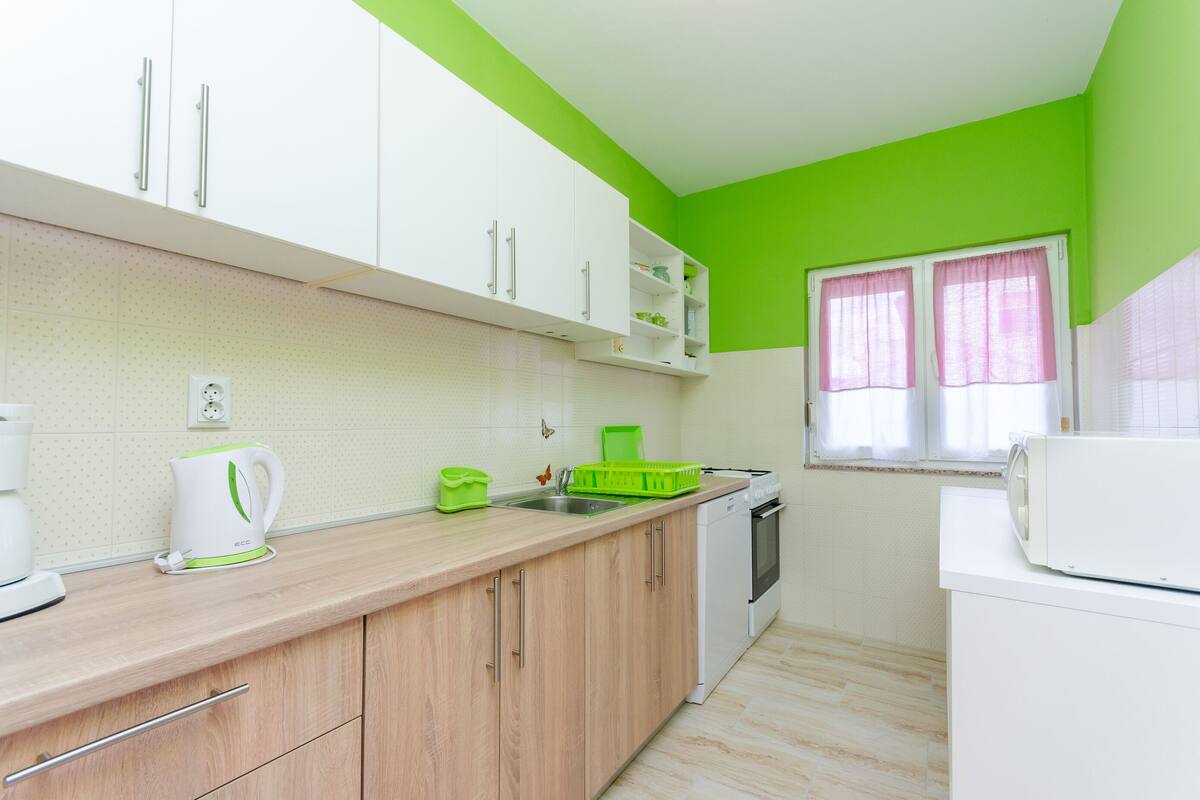 Una cucina con mobili bianchi, una parete verde e una finestra con tende rosa.