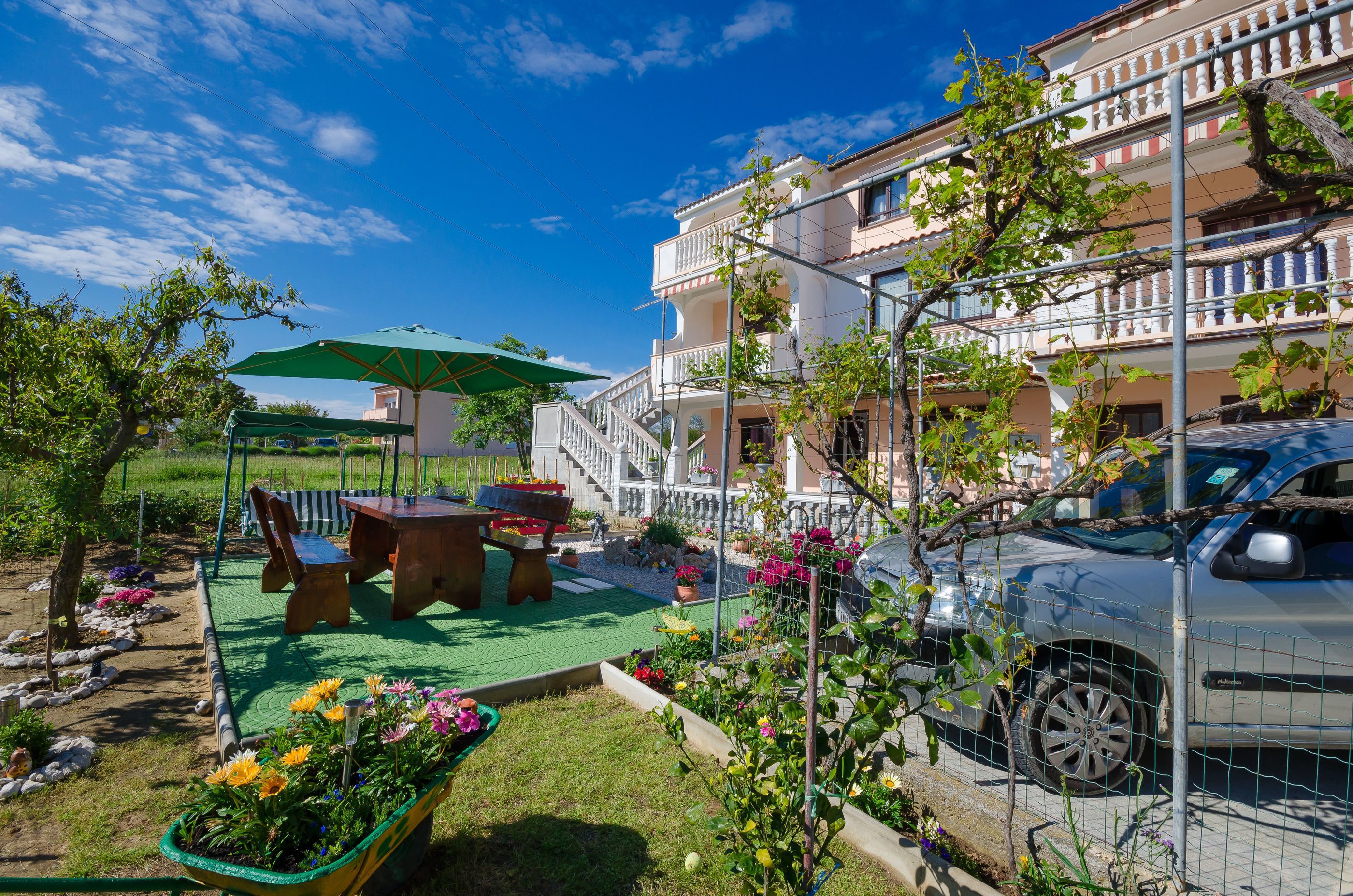 A residential building with a balcony, a car parked in front, and a garden area with a table and chairs.