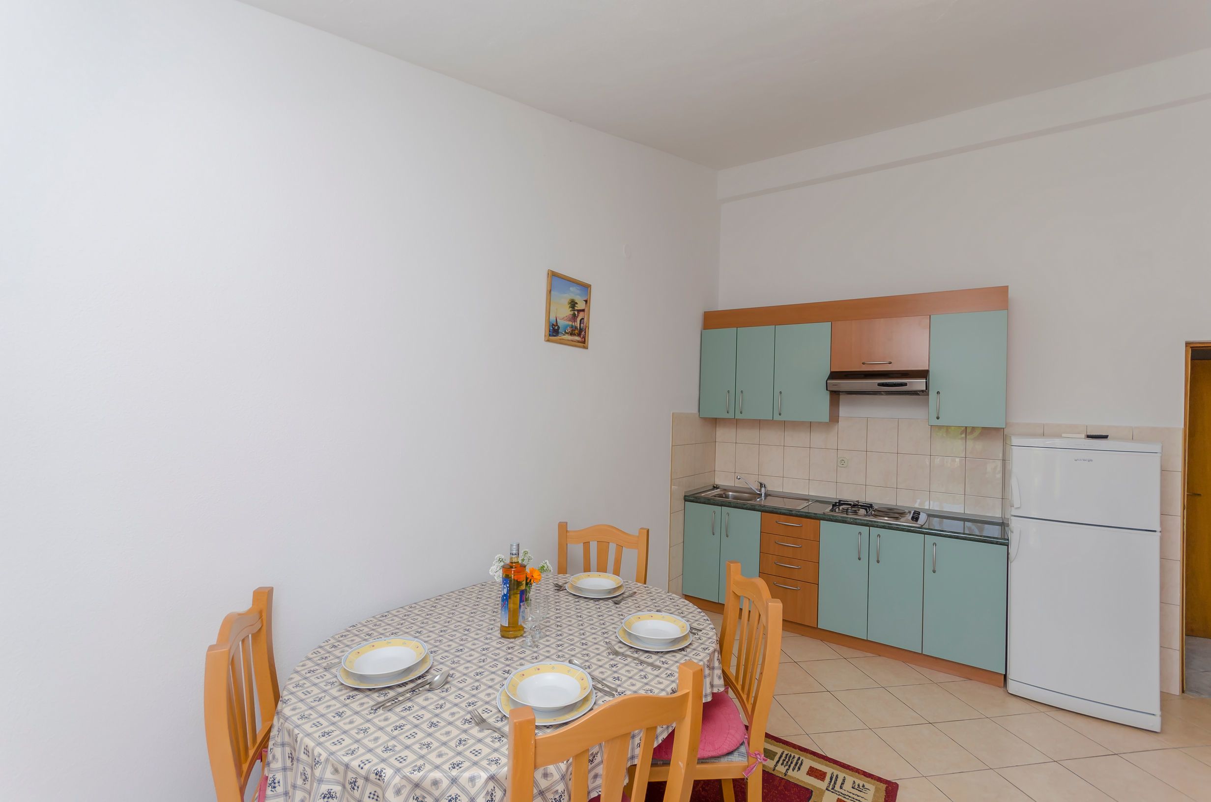 A compact kitchen with teal cabinets, a white refrigerator, and a dining table set with plates and a vase.