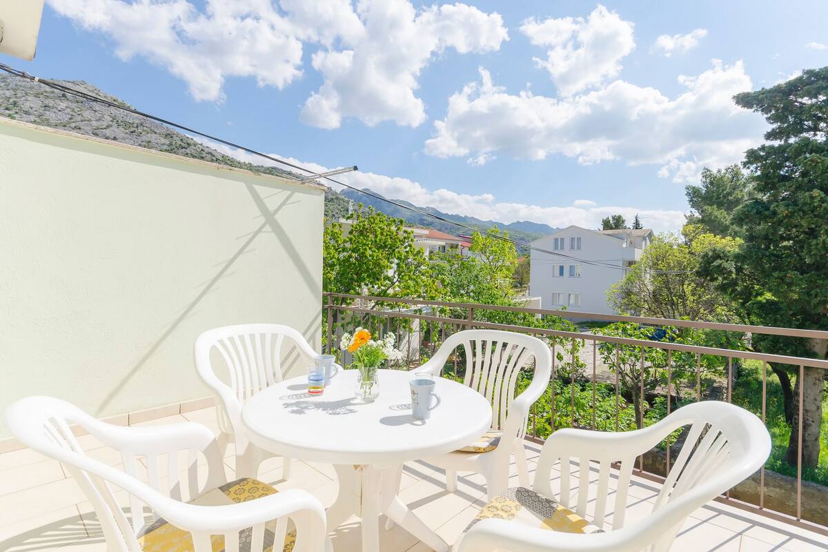 Un balcon avec une table et des chaises blanches, un petit vase avec des fleurs et une vue dégagée sur le paysage et les bâtiments environnants.
