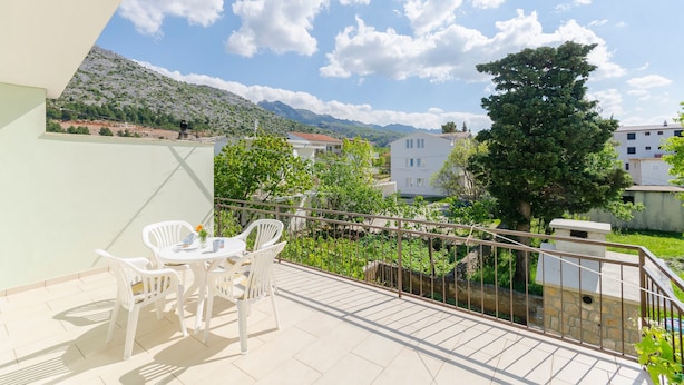 Un balcone con tavolo e sedie, affacciato su un paesaggio montuoso e edifici residenziali.