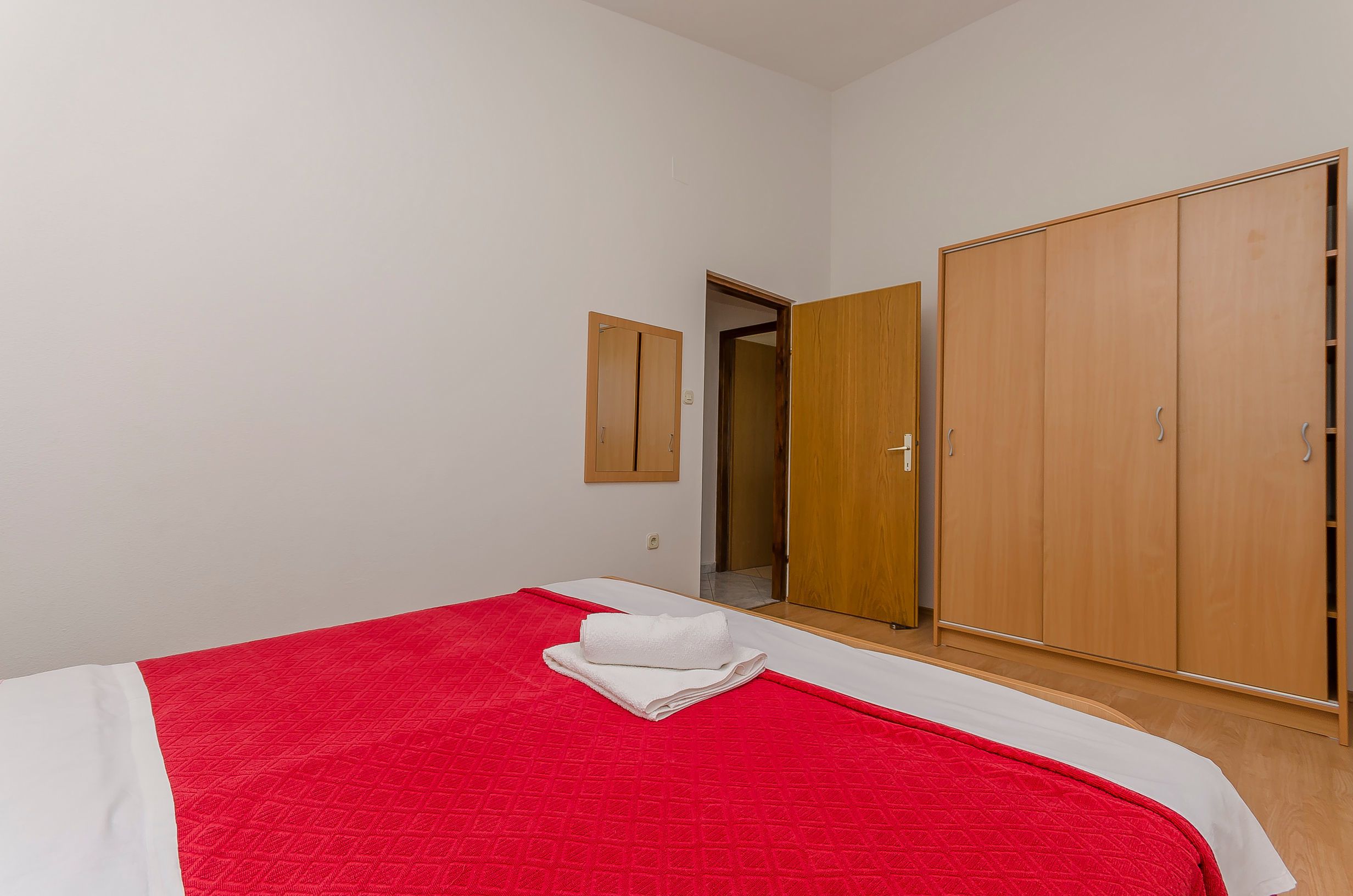 A bedroom with a bed, a red and white bedspread, a wooden wardrobe, and a mirror on the wall.