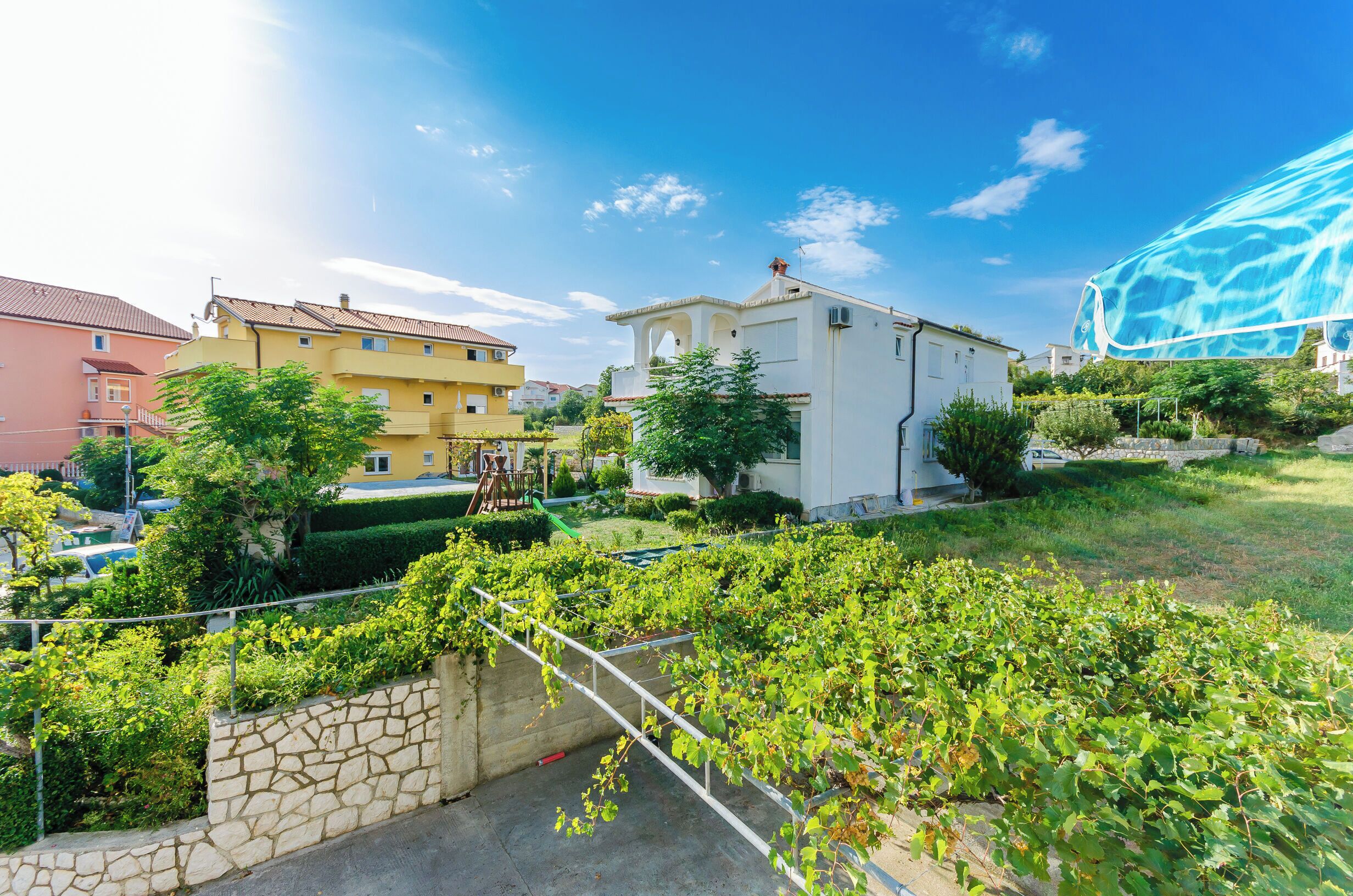 A residential area with a white building, a yellow building, and a pink building. There are green bushes and a stone wall.