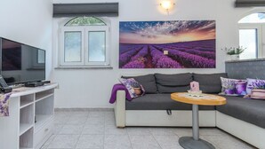 Ein Wohnzimmer mit einer Couch, einem kleinen runden Tisch, einem Fernseher und einem Fenster mit Jalousien.