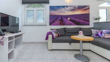 Una sala de estar con sofá, una mesita redonda, televisor y ventana con persianas.