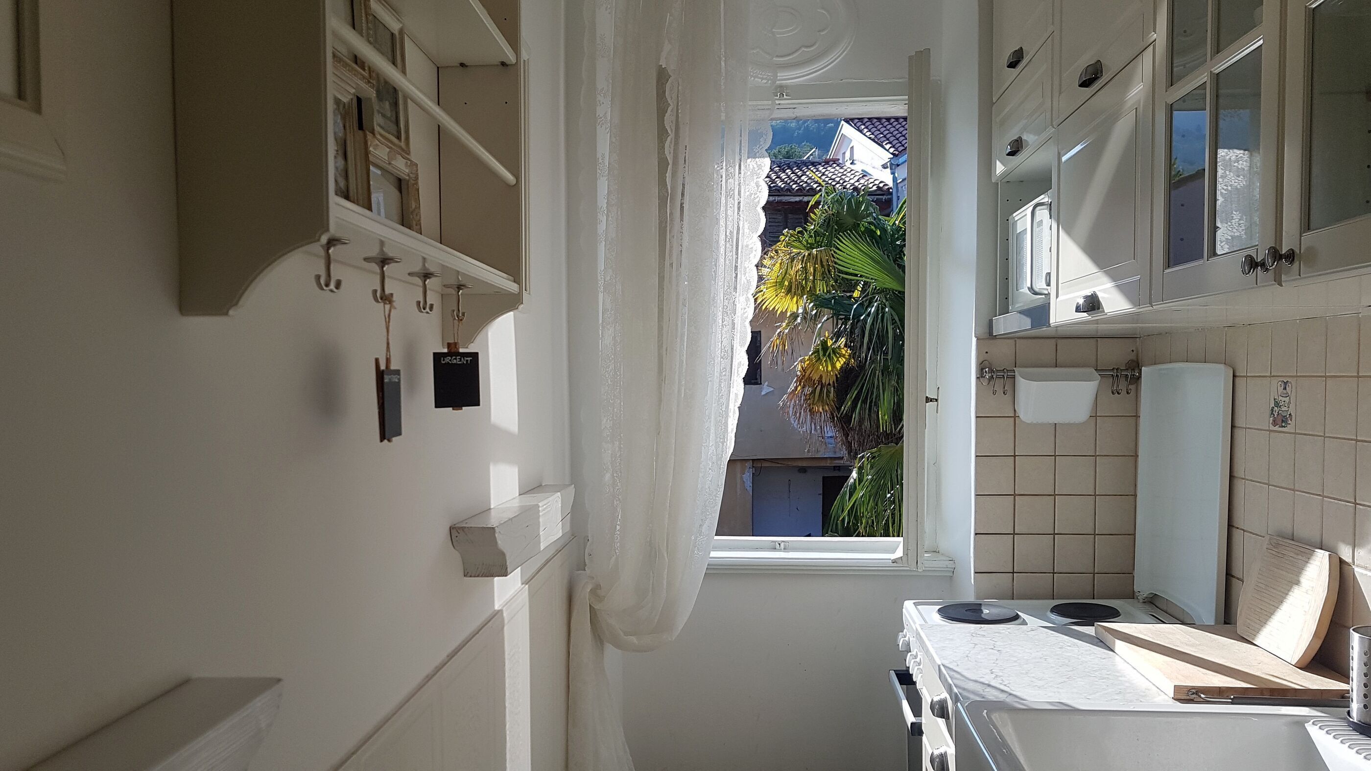 Eine schmale Küche mit weißen Schränken, einem Herd und einem Fenster, das auf einen Balkon mit Blick auf Palmen führt.